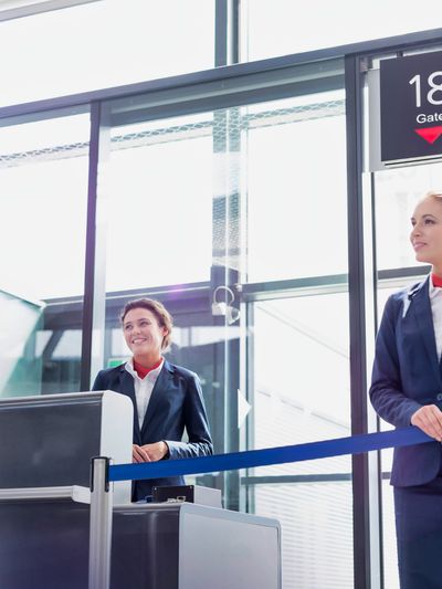 An airline gate agent stands at the gate door with tape partially closing it while another one stands at the desk.