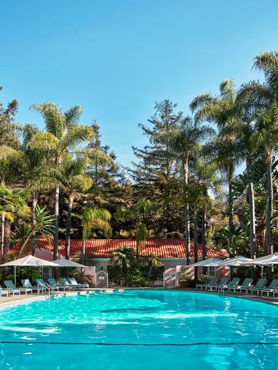 A round circular pool surrounded by pool chairs with a Mediterranean-style building behind it and towering green trees all around.