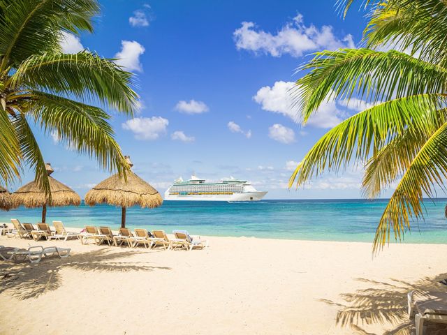 A cruise ship in the distance, as seen from a tropical beach with beach chairs, thatched umbrellas, and palm trees.