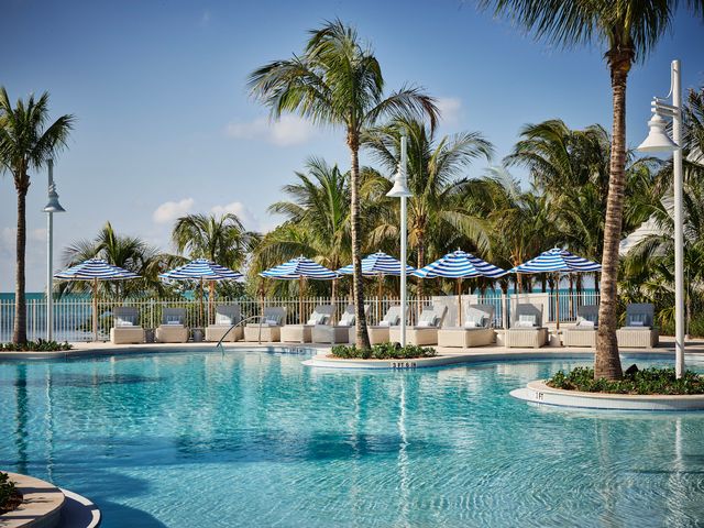 A view of a pool surrounded by palm trees and blue and white striped umbrellas with a nearby beach.