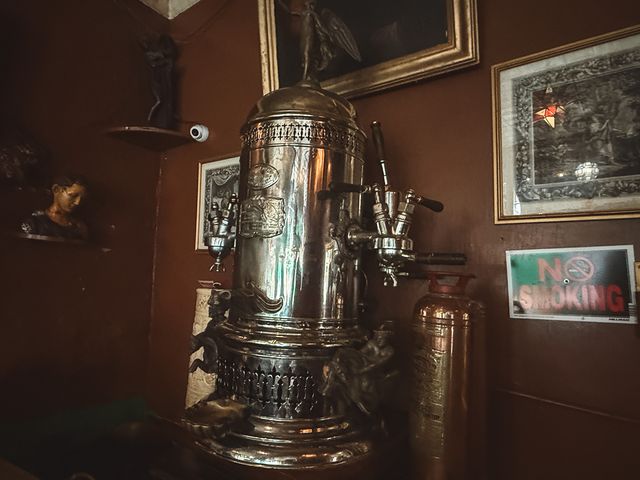 An old silver coffee machine surrounded by framed pictures.