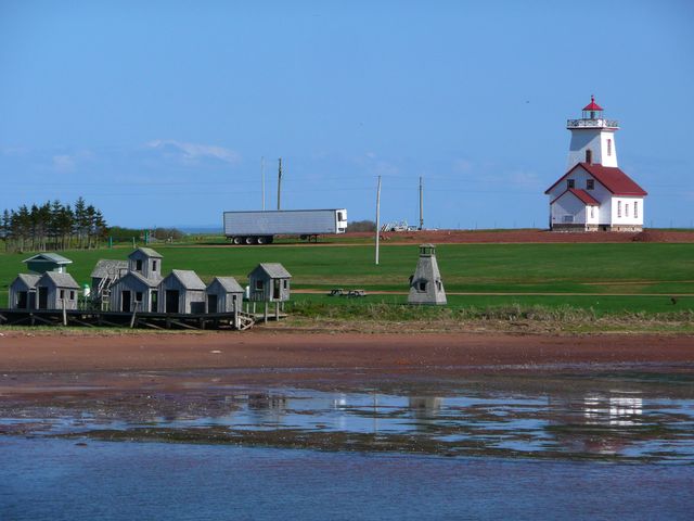 Water at low tide is in the foreground with small wooden buildings behind it, as well as a lighthouse on Prince Edward Island.