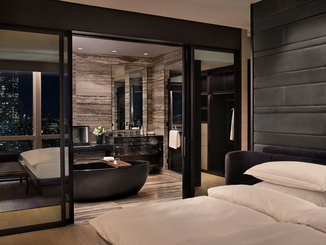 A hotel guest room with dark, black and moody color scheme looking into an open bathroom with a large black soaking tub.