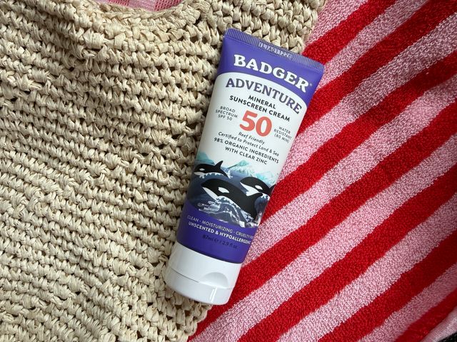 A purple and white-colored mineral sunscreen tube with orca whale pictures on top of a straw beach bag and a striped towel.