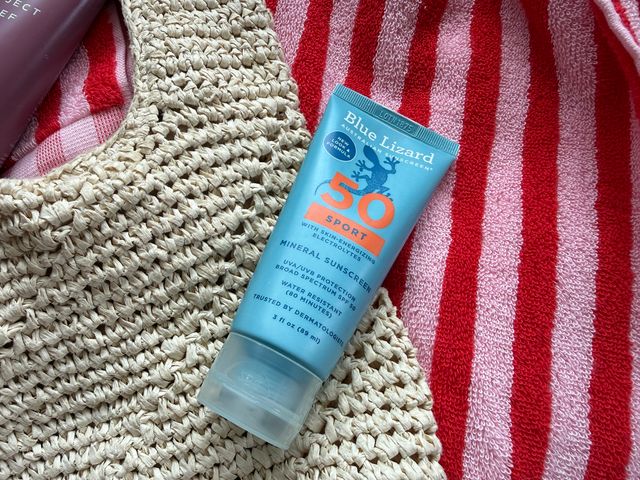 A blue mineral sunscreen tube with a lizard on it, on top of a straw beach bag and a red striped beach towel.