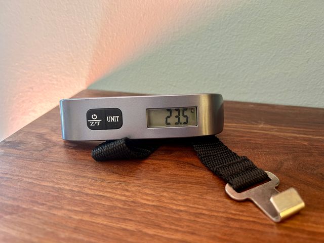 A digital luggage scale reading 23.5 sits on a wooden table.