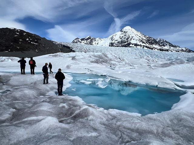 A group of people are seen standing next to a blue pool of water on a glacier in Alaska.