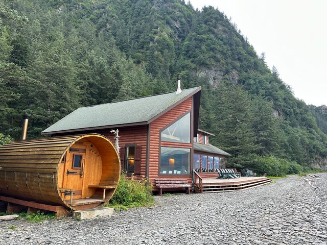 A log-style cabin next to a barrel sauna and pine trees.