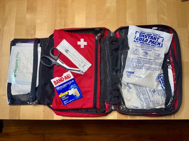 A red cross first aid kit open across a table with scissors, bandages, and a cold pack visible.