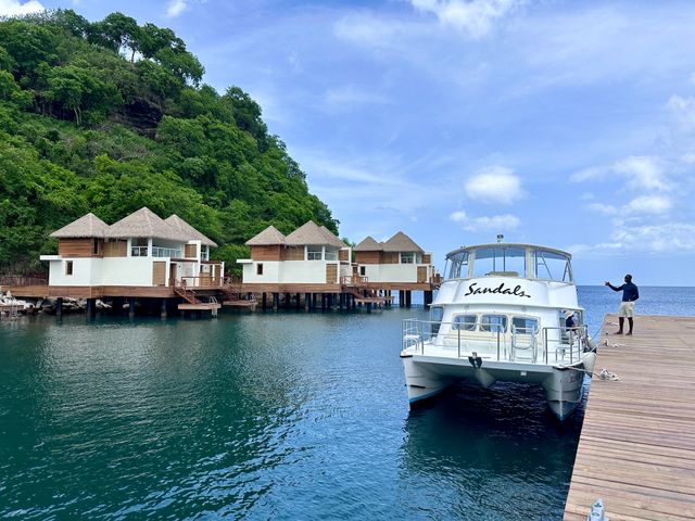 Overwater rooms at a Sandals Resort are seen between a green hillside and water next to a dock with a boat.