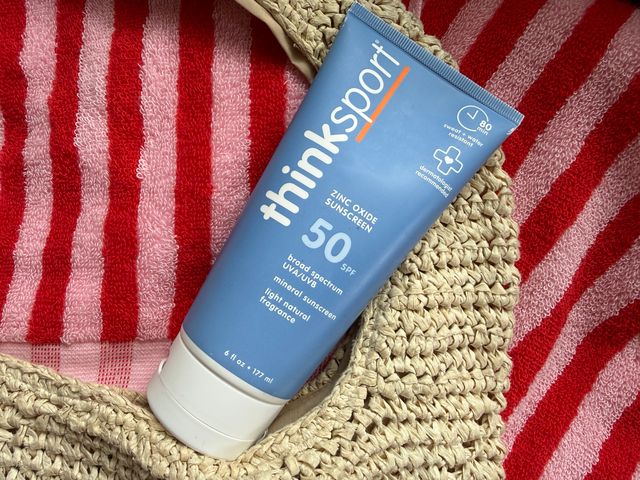 A blue Thinksport mineral sunscreen tube with a 50 SPF label on it on top of a straw beach bag and a red striped beach towel.
