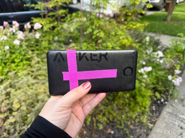 A hand holding an Anker branded black power bank with pink tape on it in front of a bush.