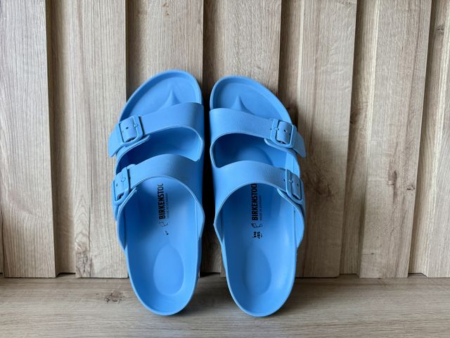 A pair of electric blue Birkenstock sandals leaning on a wood wall on wood floors.