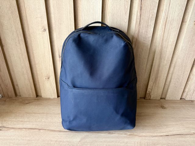 A small dayback backpack leaning against a wood wall on wood floors.