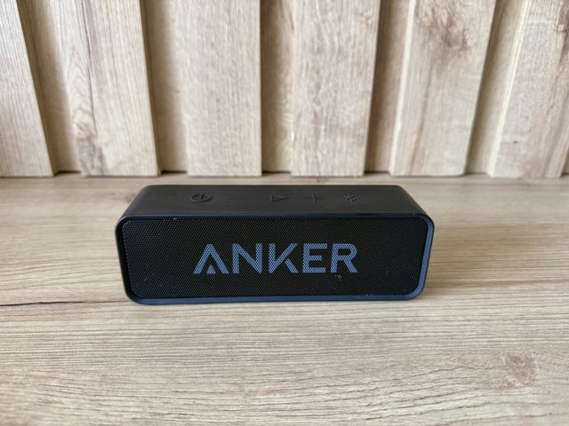 A small black portable speaker with the word Anker on it, against a wood wall on wood floors.