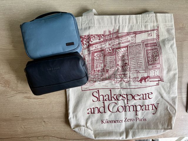 A small black pouch next to a small blue bouch and a Shakespeare and Company canvas tote bag, all on wood floors.