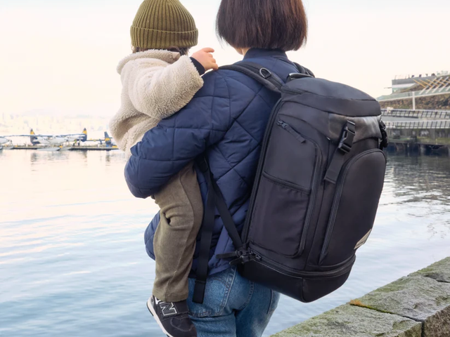 A woman is seen from behind wearing a backpack while holding her baby and looking out at a waterfront landscape.