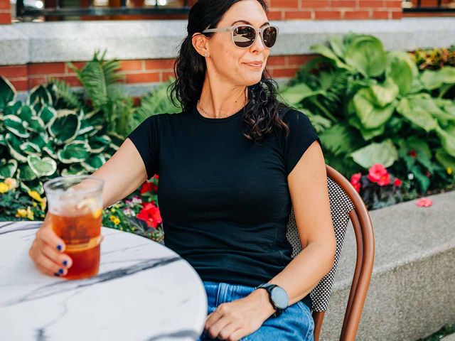 A woman wearing sunglasses and a black t-shirt at a sidewalk table outside with a drink.