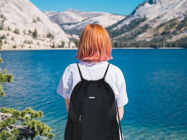 A woman with strawberry hair sitting with her back visible, facing a body of water, wearing a black drawstring nylon backpack.