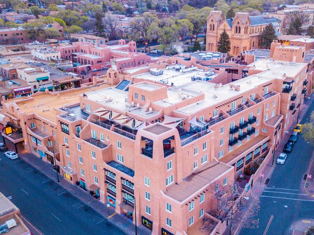 The exterior of La Fonda on the Plaza on a city corner with red stucco walls surrounded by city buildings and trees, as seen from above.