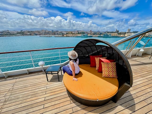 A woman sitting on an orange daybed on a cruise deck looking out at port on a Windstar Cruise in the Canary Islands.