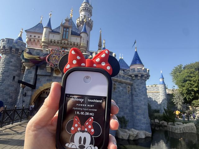 A hand holds out a bottle of hand sanitizer in a Minnie Mouse case in front of the castle at Disneyland.