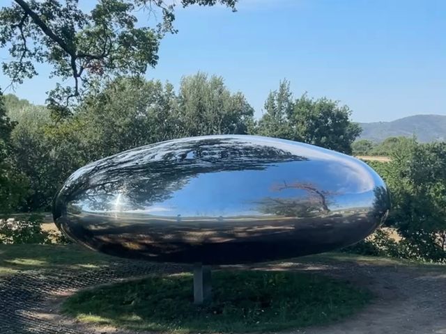 A large silver cylindrical art installation next to green trees and vineyard fields.