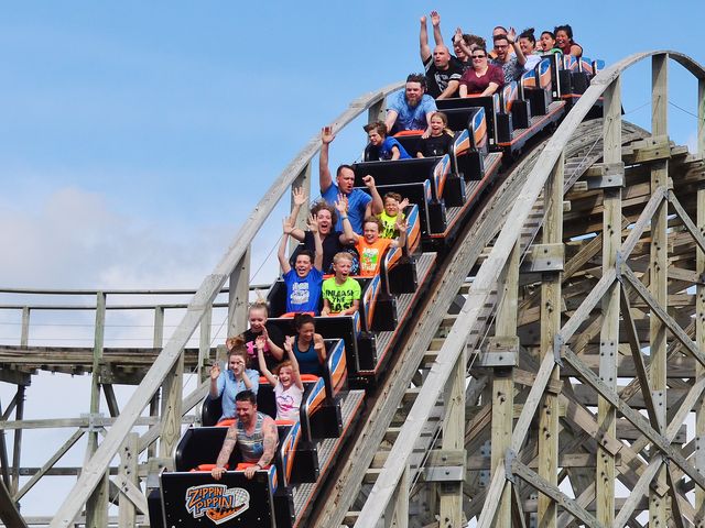 A mixed crowd of children and adults ride on a wooden rollercoaster with their hands in the air