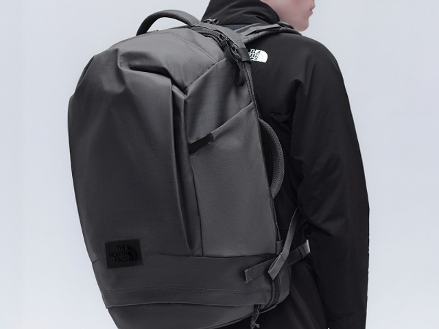 A man wearing a black jacket, black sunglasses, and a black backpack against a white background.