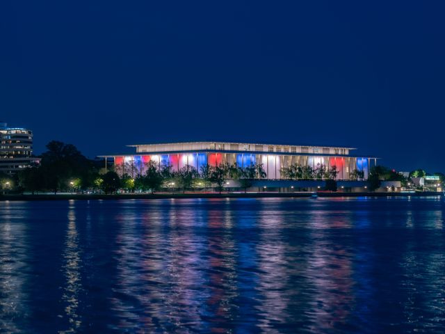 The exterior of the Kennedy Center at night lit up with red, white, and blue colors next to the Potomac River.