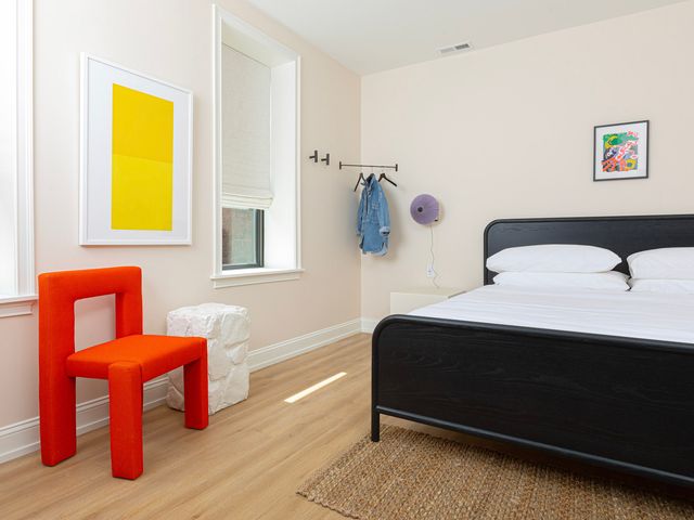 A room at the Yowie Hotel in Philadelphia with a bright red chair, framed art, a bed, and a corner hanger with clothes.