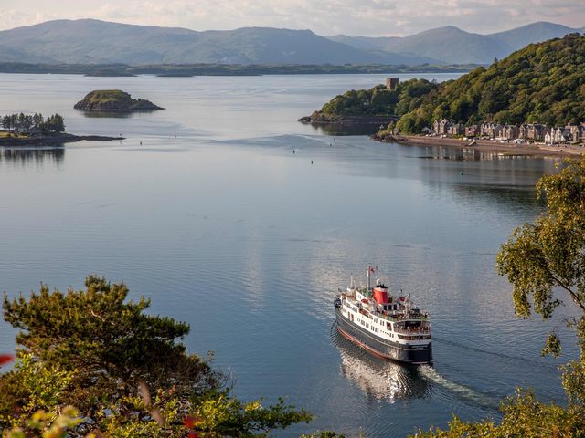 A ferry is seen on the water next to rugged shores with trees and stately buildings on the coast.