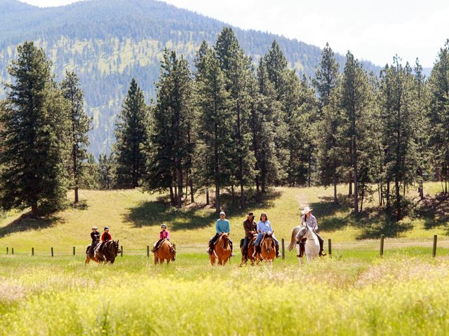 A family with members of all ages is seen riding horses in a field next to pine trees and mountains at Paws Up Montana.
