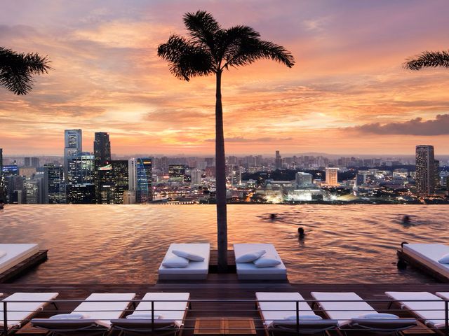 A rooftop infinity pool with in-water beds next to palm trees and sunset city views of Singapore at Marina Bay.