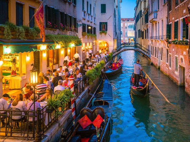 Three gondolas in a canal in Venice next to a crowded restaurant patio.