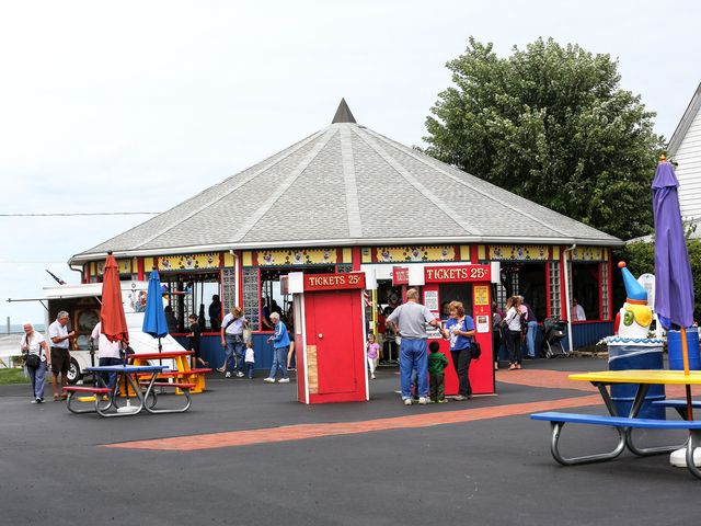 Amusement park guest mill around the covered carousel on a blacktop near an old-school red ticket booth
