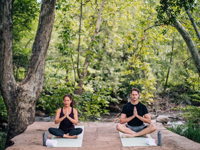 A man and a woman sitting on mats surrounded by lush green trees in a yoga class.