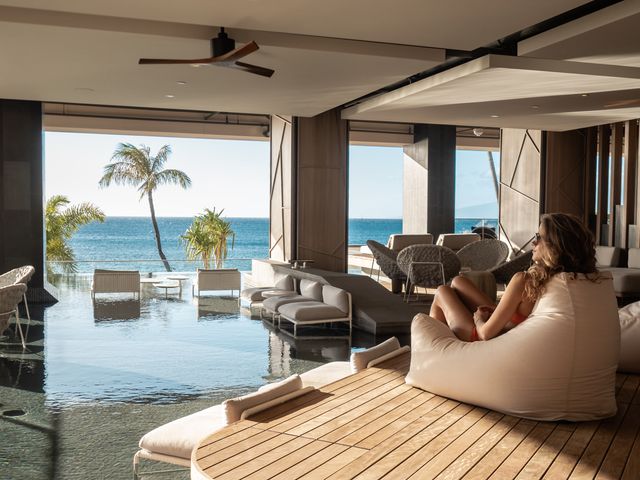 A woman sits on a plush chair on a deck next to an indoor pool that's also outdoor with open-air walls overlooking the ocean.
