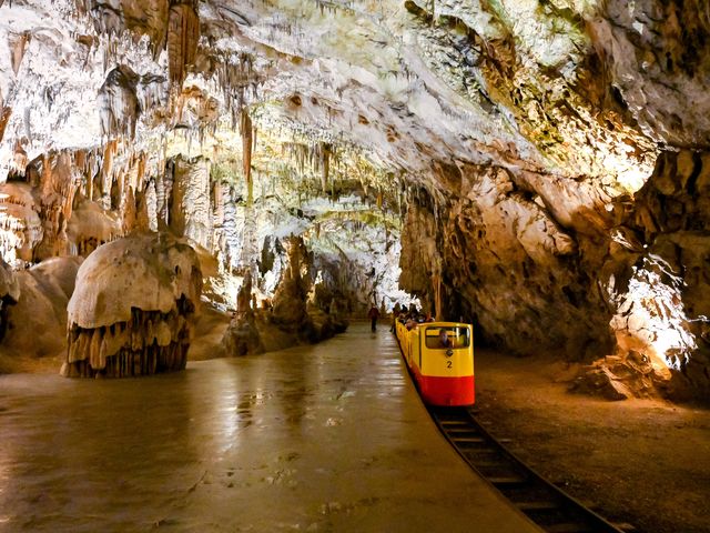 An underground yellow and red tourist train in Postojna cave, Slovenia surrounded by stalactite and stalagmite formations.