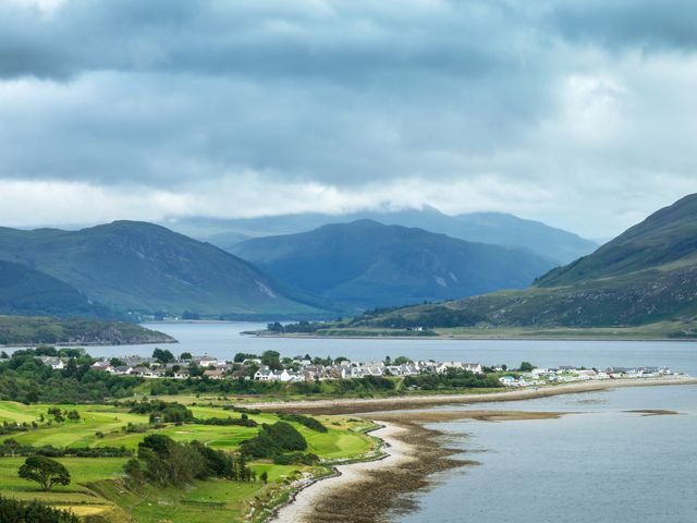 A sweeping view over the coastal area of Ullapool, Scotland with a small coastal town and green mountains.