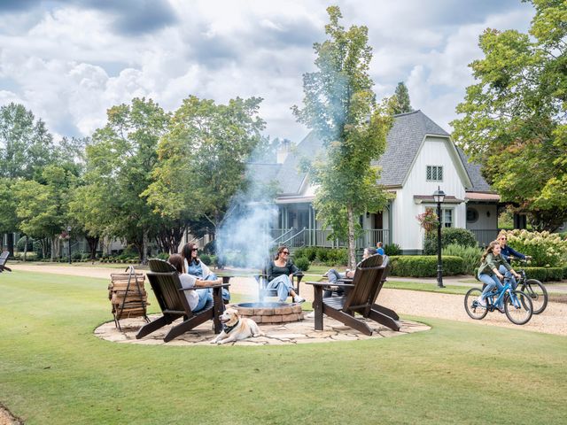 A group of people sit around a fire pit at Barnsely Resort as bike riders pass by next to a building and green trees.