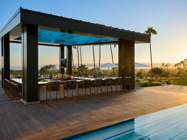 A large wraparound bar shaded by a screen projecting images of fish next to a pool, at sunset.
