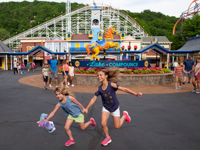 Two smiling young girls run in front of the entrance sign to Lake Compounce park with a white rollercoaster behind them.