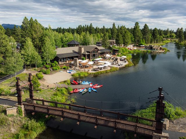 A body of water with kayaks at a dock, a bridge going over the water, and a building surrounded by trees as seen from above at Sunriver Resort.