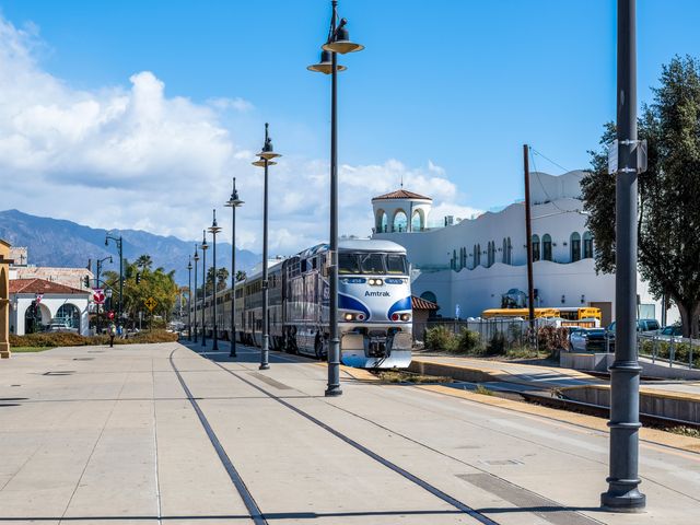 An Amtrak Pacific Surfliner train at Santa Barbara Station surrounded by Mission-style architecture, trees, and mountains.