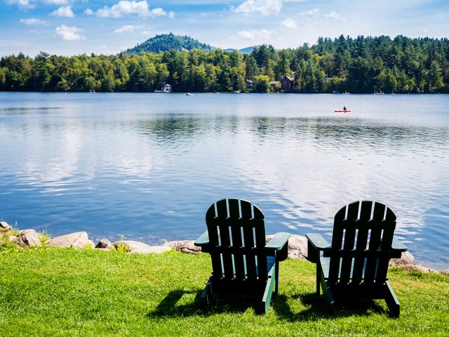 Two empty adirondack chairs overlooking Mirror Lake in Lake Placid, with a kayaker in the distance.