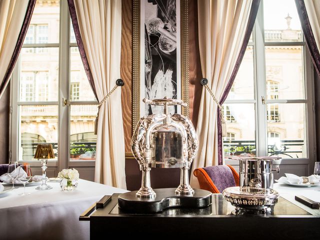 A fancy hotel dining room with large windows framed with curtains, plush purple and orange chairs, and a tray table on wheels with silver serveware.
