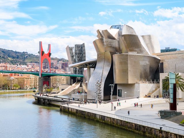 The modern, futuristic exterior of the Guggenheim Bilbao museum on a sunny day with a bridge behind it.