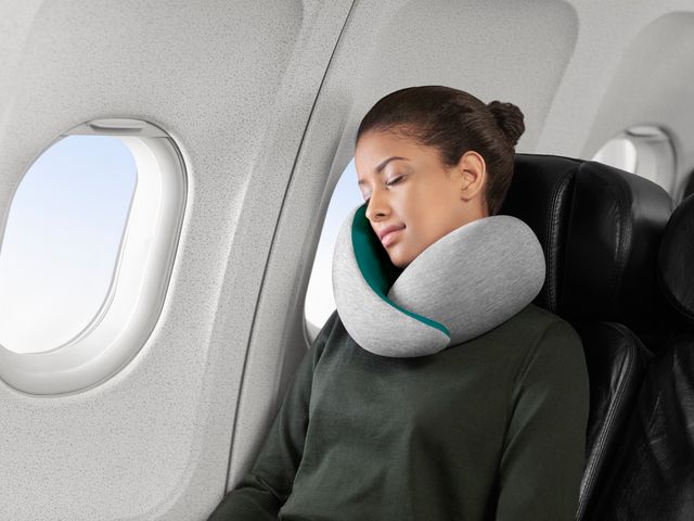 A woman sleeps in the window seat of an airplane while wearing a supportive neck pillow.