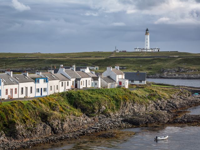 A row of white buildings on a rugged coast in Islay with a lighthouse in the background.